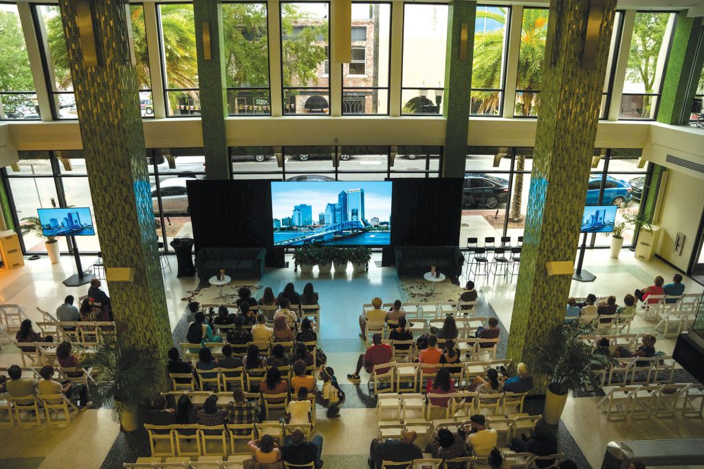 photo of people in seating watching video on large screen in large open room
