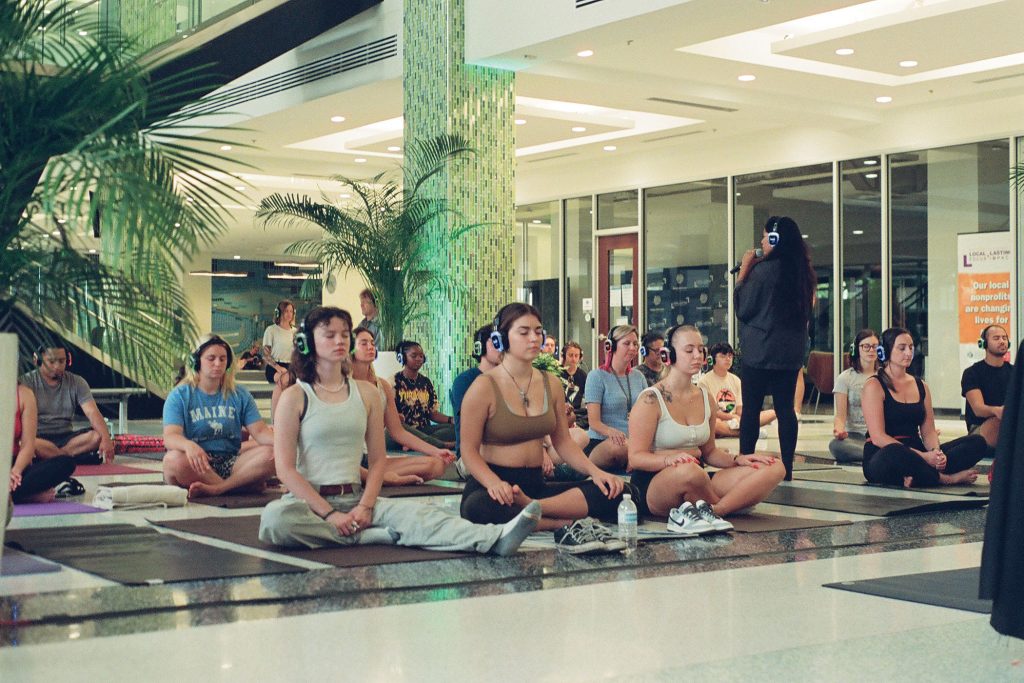 photo of people sitting cross-legged on floor with eyes closed while a woman walks among them and talks into microphone