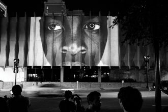 black and white photo of building exterior with image of black male face projected onto building